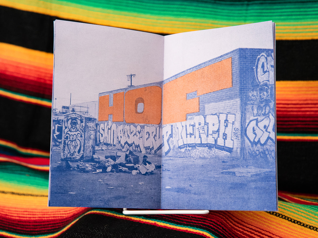 A photography zine with a blue cover featuring orange text and a black and white image of graffiti on a wall. The zine is placed on a surface with a multicolored pattern.