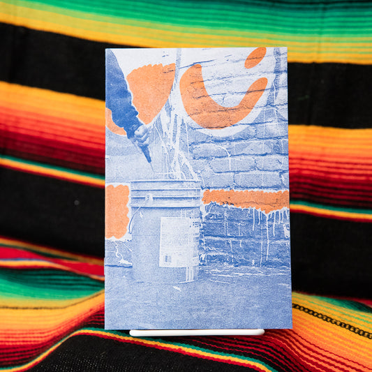 A photography zine with a blue cover featuring orange text and a black and white image of graffiti on a wall. The zine is placed on a surface with a multicolored pattern.